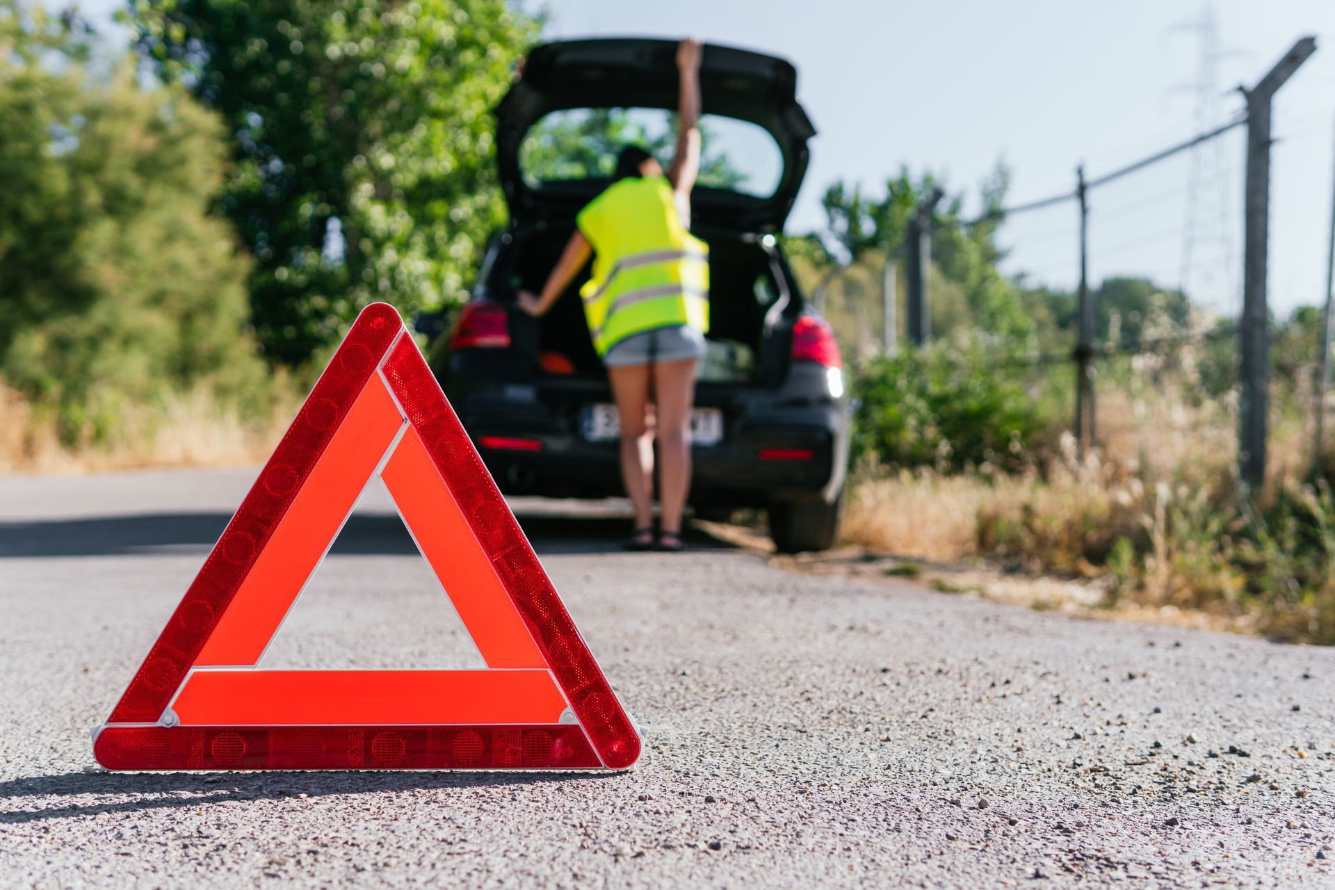 Triangle de signalisation rouge en bord de route, près d'une personne inspectant une voiture en panne dont le coffre est ouvert.