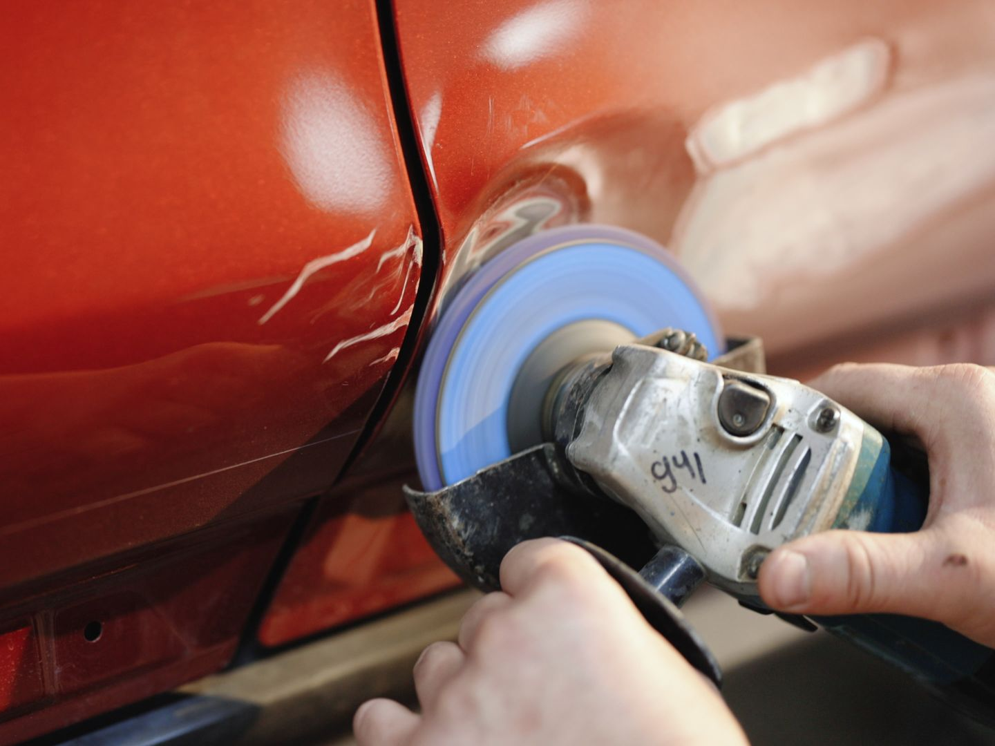 Main utilisant un outil de polissage sur une portière de voiture rouge, lustrant la peinture près d'un joint.