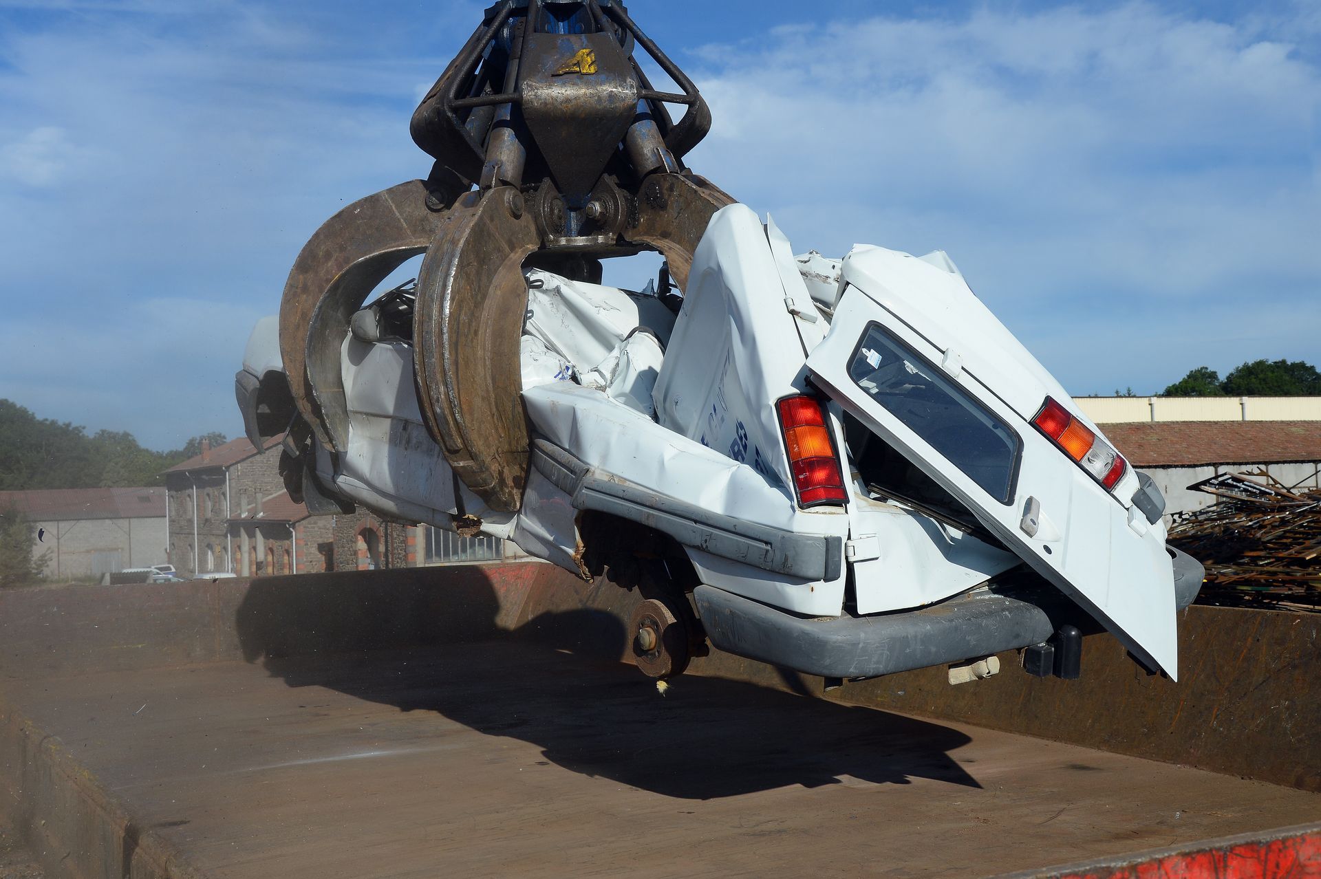 Une camionnette blanche est écrasée et soulevée par une grande pince métallique dans une cour extérieure poussiéreuse.