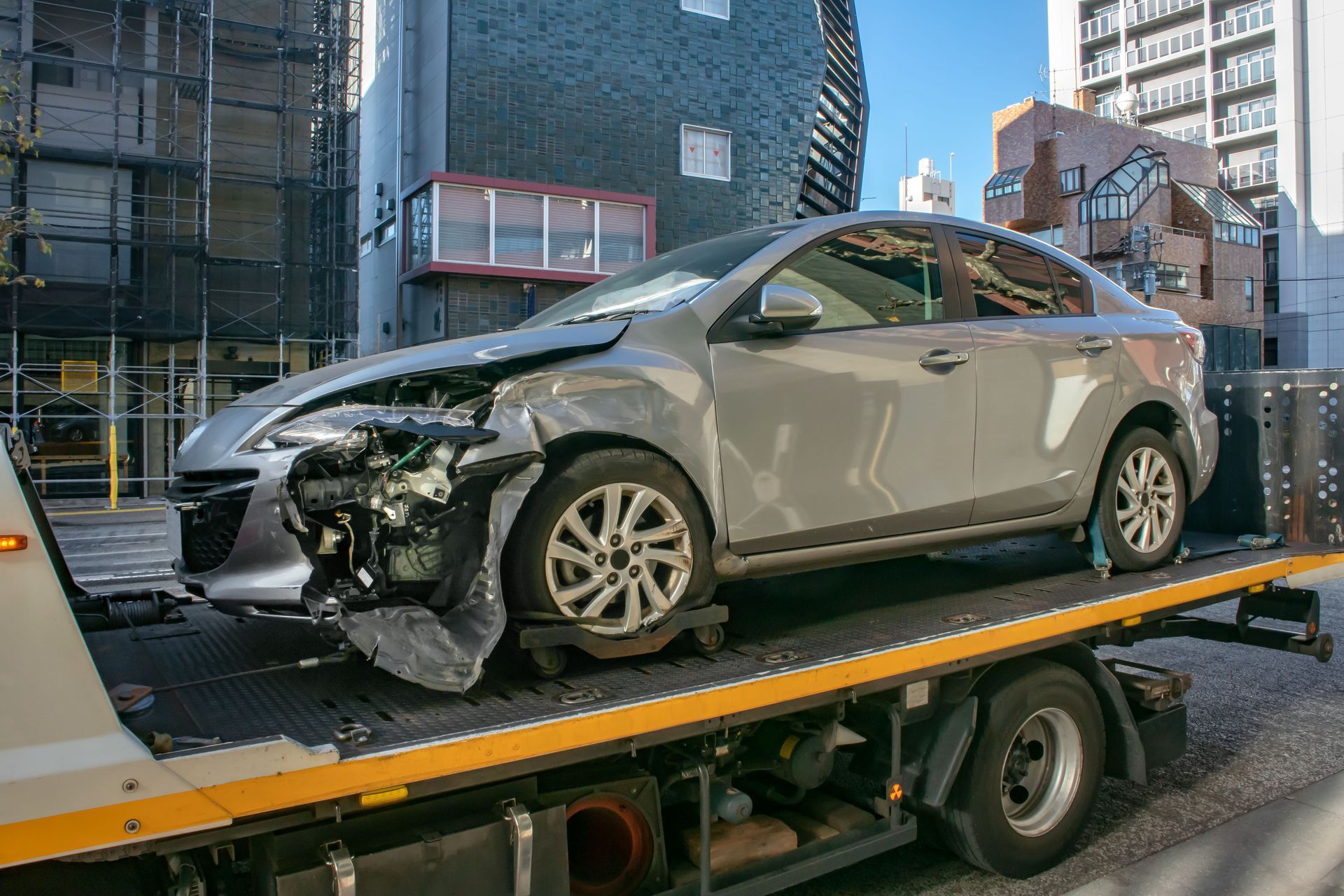 Voiture argentée endommagée sur une dépanneuse dans une rue de la ville, pièces de carrosserie manquantes.
