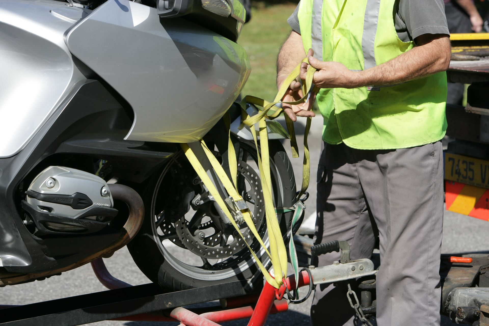 Un ouvrier portant un gilet fluo attache une moto à un attelage de remorque rouge.