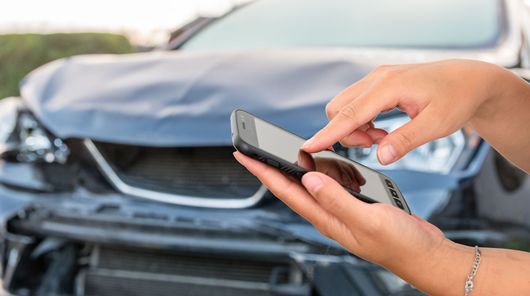 Des mains utilisent un smartphone pour contacter une dépanneuse après un accident, voiture endommagée en arrière-plan.
