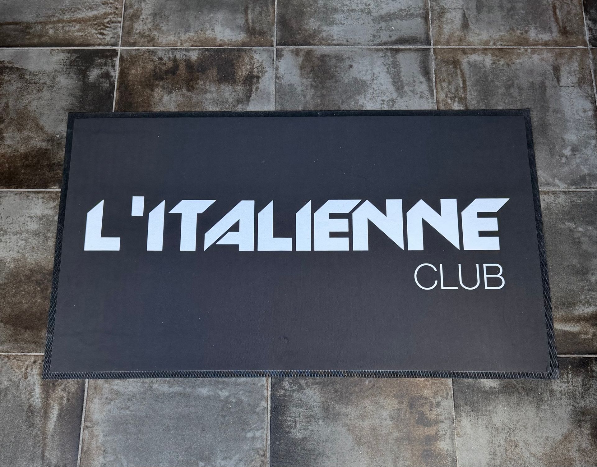 Paillasson noir avec l'inscription blanche « L'ITALIENNE CLUB » sur un sol carrelé.
