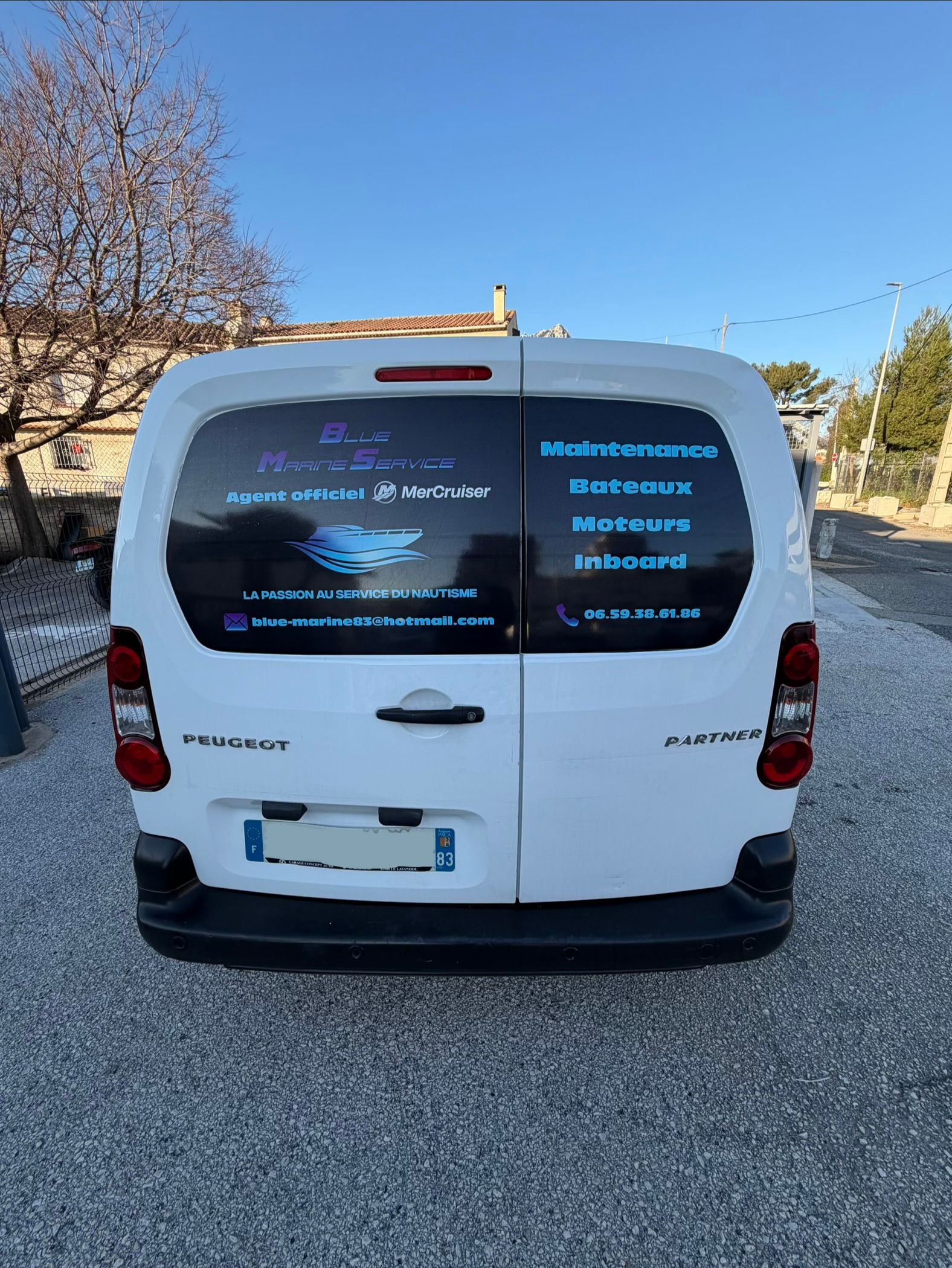 Une camionnette blanche avec des inscriptions bleues et noires sur la lunette arrière est garée sur une allée de gravier.