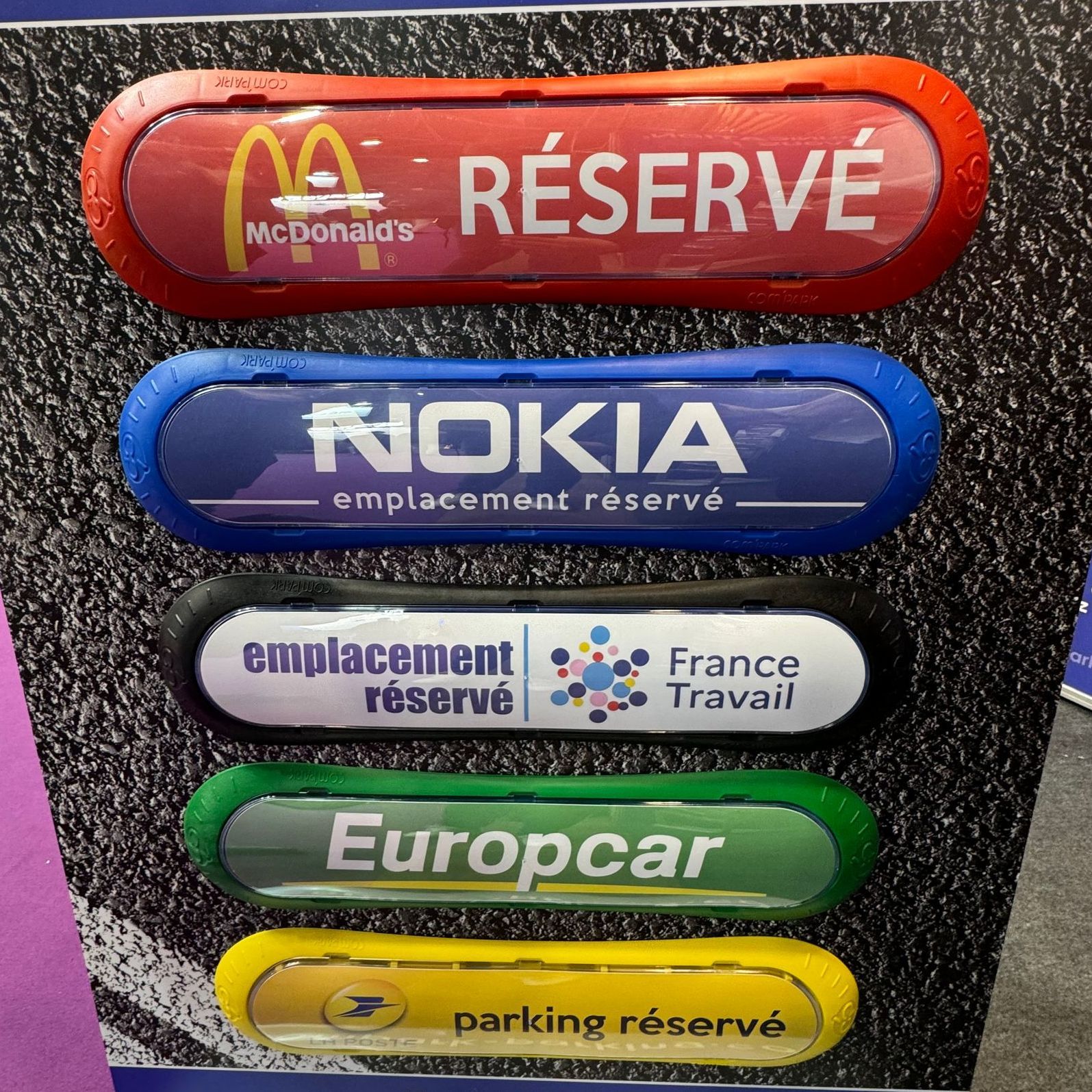 Cinq panneaux de stationnement de couleurs différentes, ornés des logos des entreprises : McDonald's, Nokia, France Travail, Europcar et La Poste. Tous comportent la mention « Stationnement réservé ».