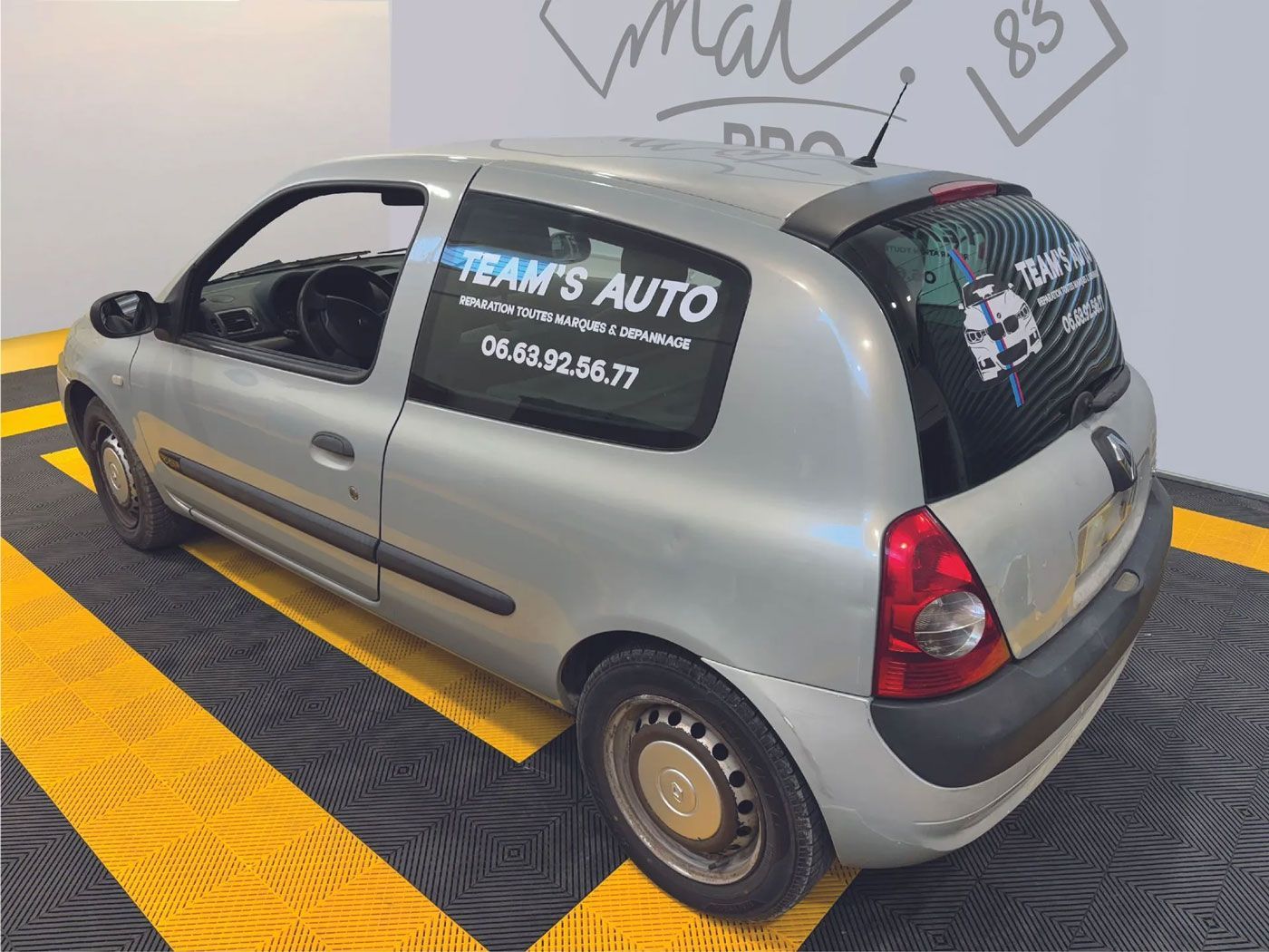 Une Renault Clio argentée, arborant une publicité de Team's Auto, est garée sur une surface à rayures jaunes et noires.