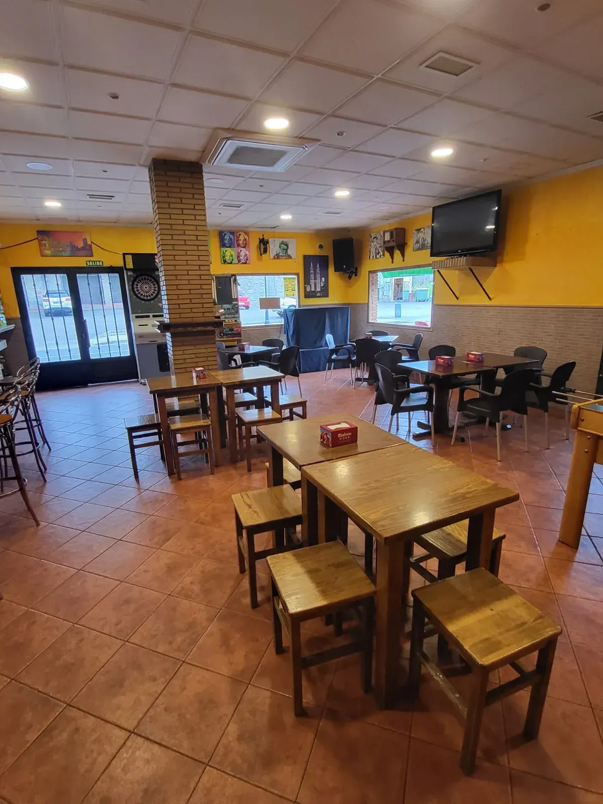 Vista interior de un bar con mesas de madera, taburetes y paredes amarillas. Hay un televisor colgado en la pared.