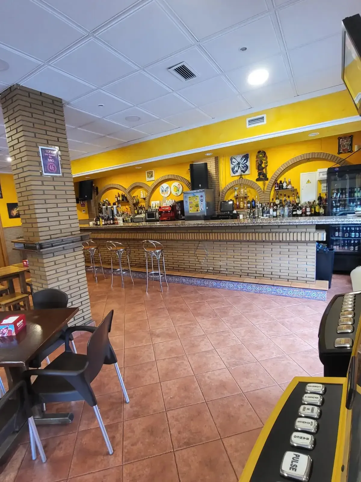Interior de un bar con fachada de ladrillo, paredes amarillas, suelo de baldosas y una larga barra de madera.