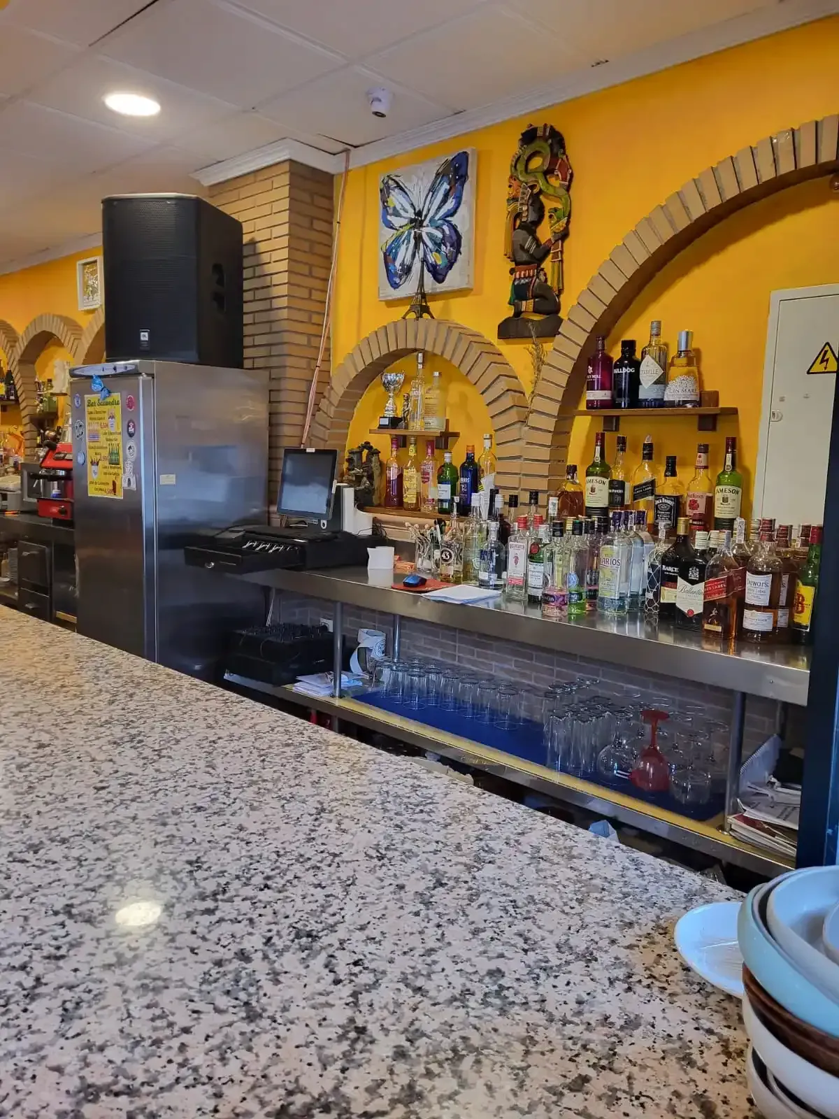 Interior de bar con encimera de mármol, barra de acero inoxidable y botellas en estantes bajo aberturas arqueadas.