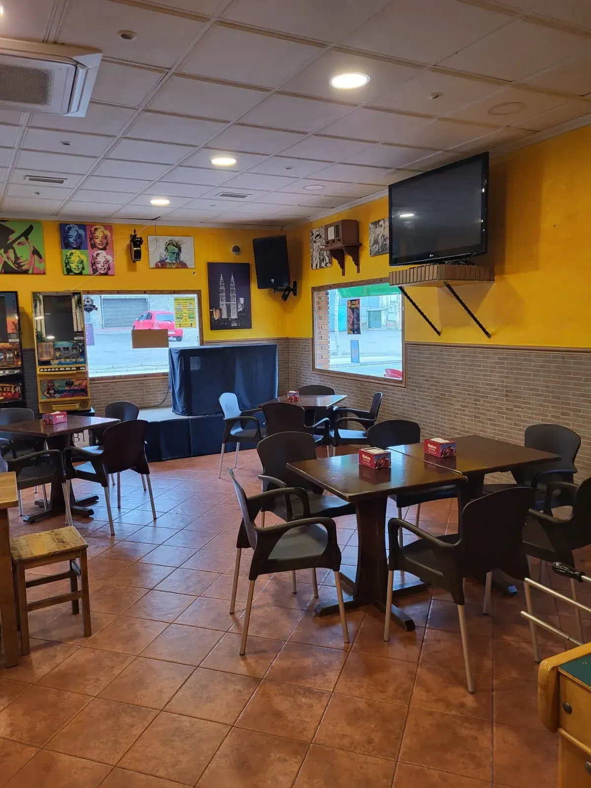 Interior de una cafetería con mesas, sillas y un televisor colgado. Paredes amarillas, suelo de baldosas y ventanas.