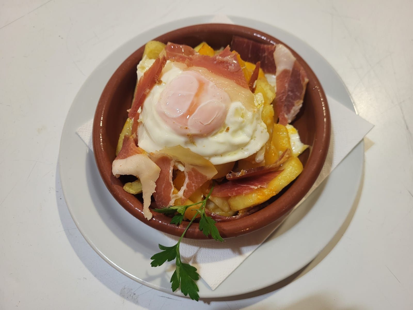 Plato de patatas fritas, cubierto con un huevo frito y jamón, en un recipiente marrón.