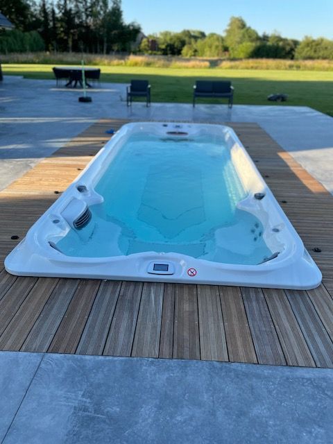 Un long spa de nage blanc rempli d'eau, installé sur une terrasse en bois, avec des sièges extérieurs et des arbres en arrière-plan.