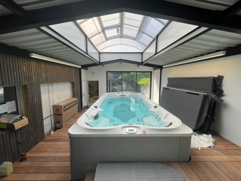 Vue intérieure d'un spa de nage rectangulaire, sous une verrière. Sol et murs en bois ; murs blancs.