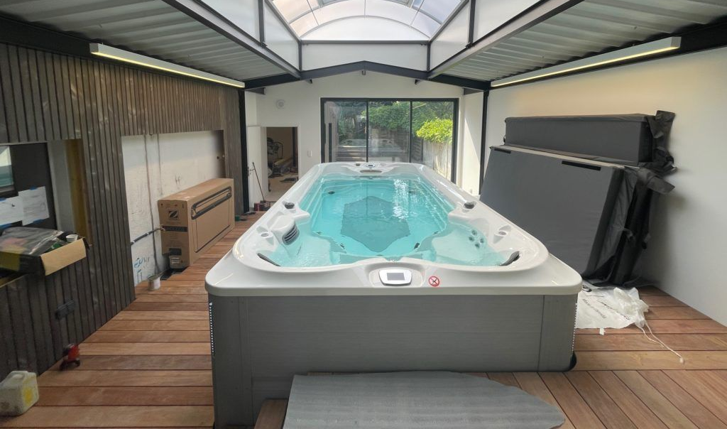 Spa de nage intérieur, blanc et gris avec eau bleue, dans une pièce avec parquet et puits de lumière.
