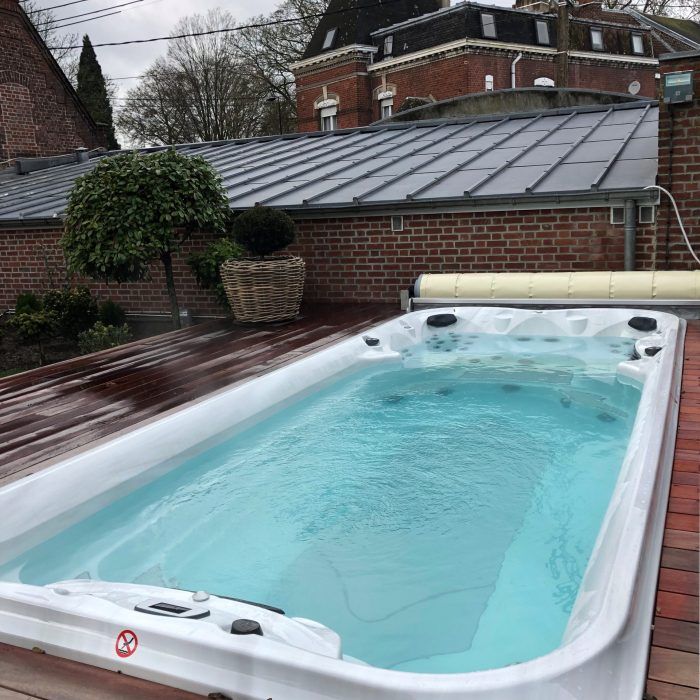 Spa de nage rectangulaire sur une terrasse en bois, rempli d'eau claire, adossé à des bâtiments en briques.