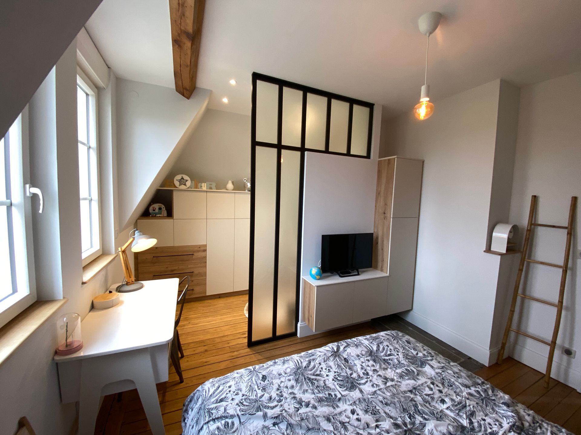 Chambre avec plafond mansardé, bureau, lit, télévision et paravent décoratif.