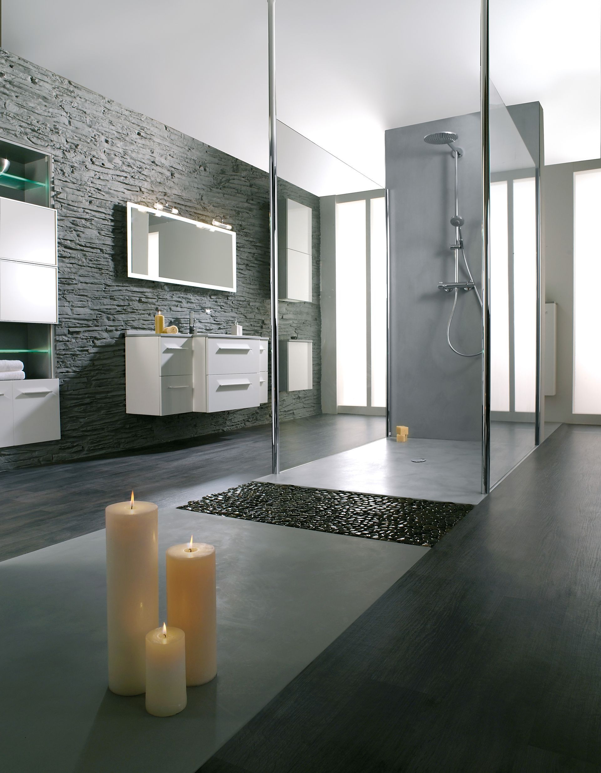 Salle de bains moderne avec mur en pierre gris foncé, douche en verre, meubles blancs et bougies allumées.