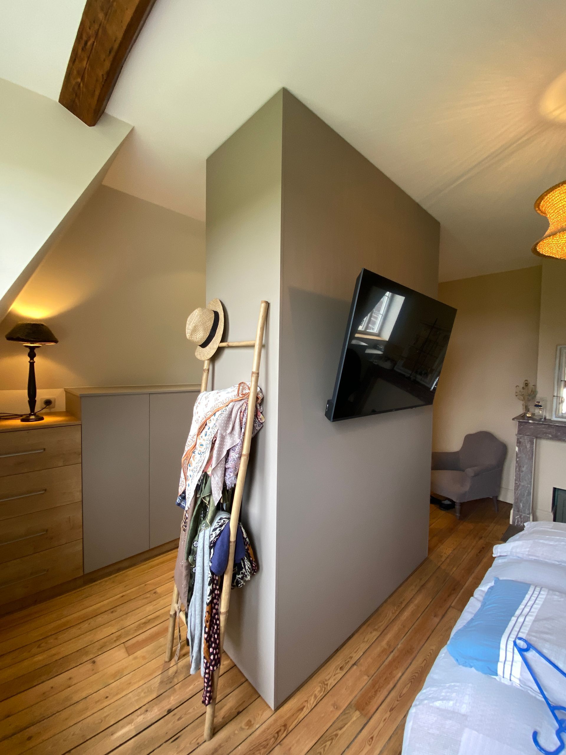 Intérieur d'une chambre avec télévision sur un mur gris, un portant à vêtements en forme d'échelle en bois et un lit.