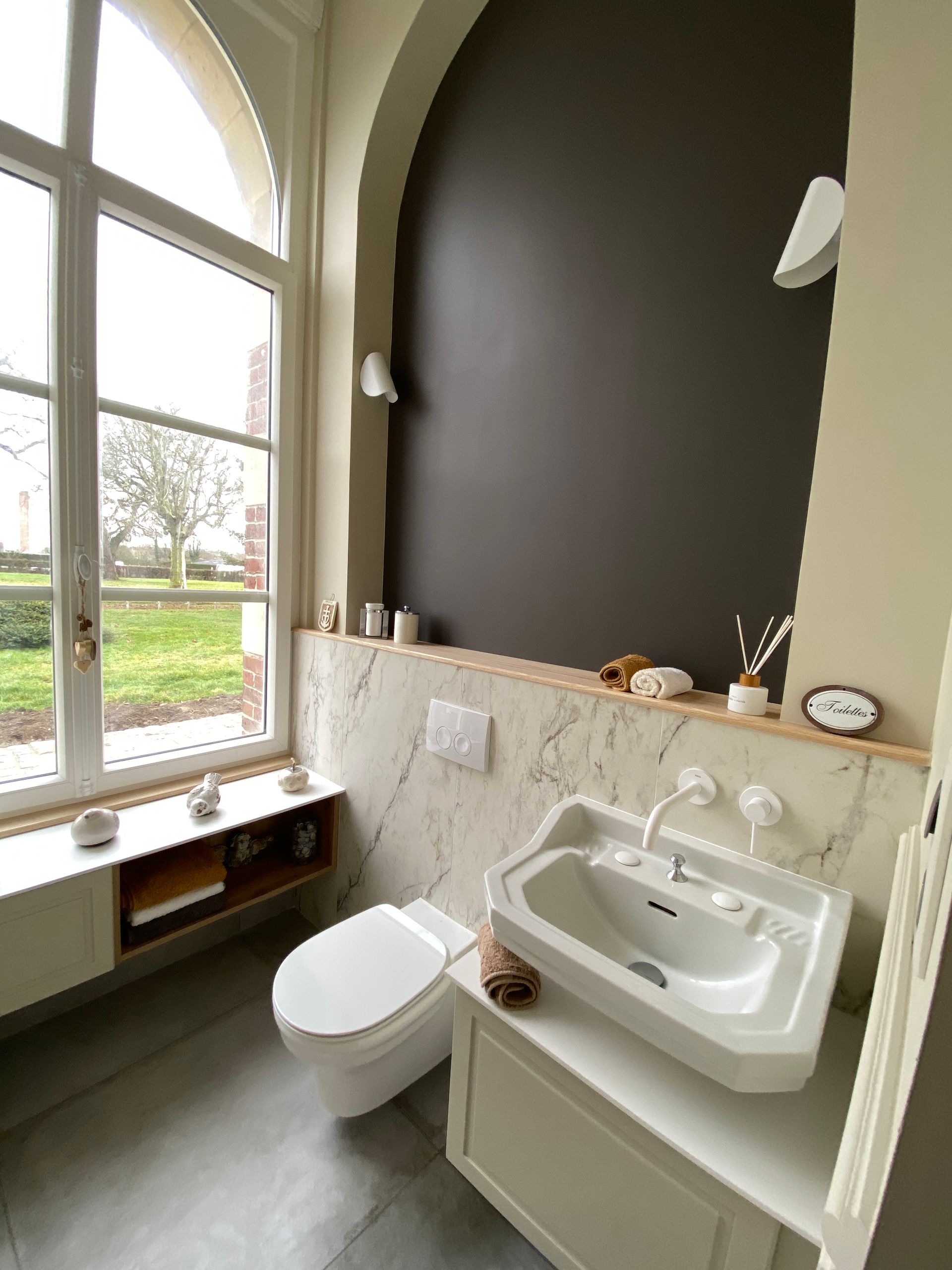 Salle de bains avec toilettes et lavabo blancs, mur en marbre, mur d'accent foncé et grande fenêtre.