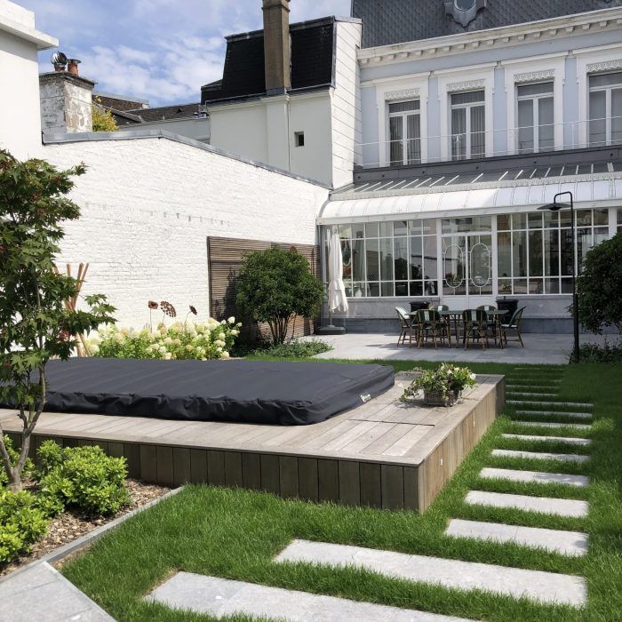 Un jardin avec un jacuzzi sur une terrasse en bois, un chemin de pierre et une maison blanche.
