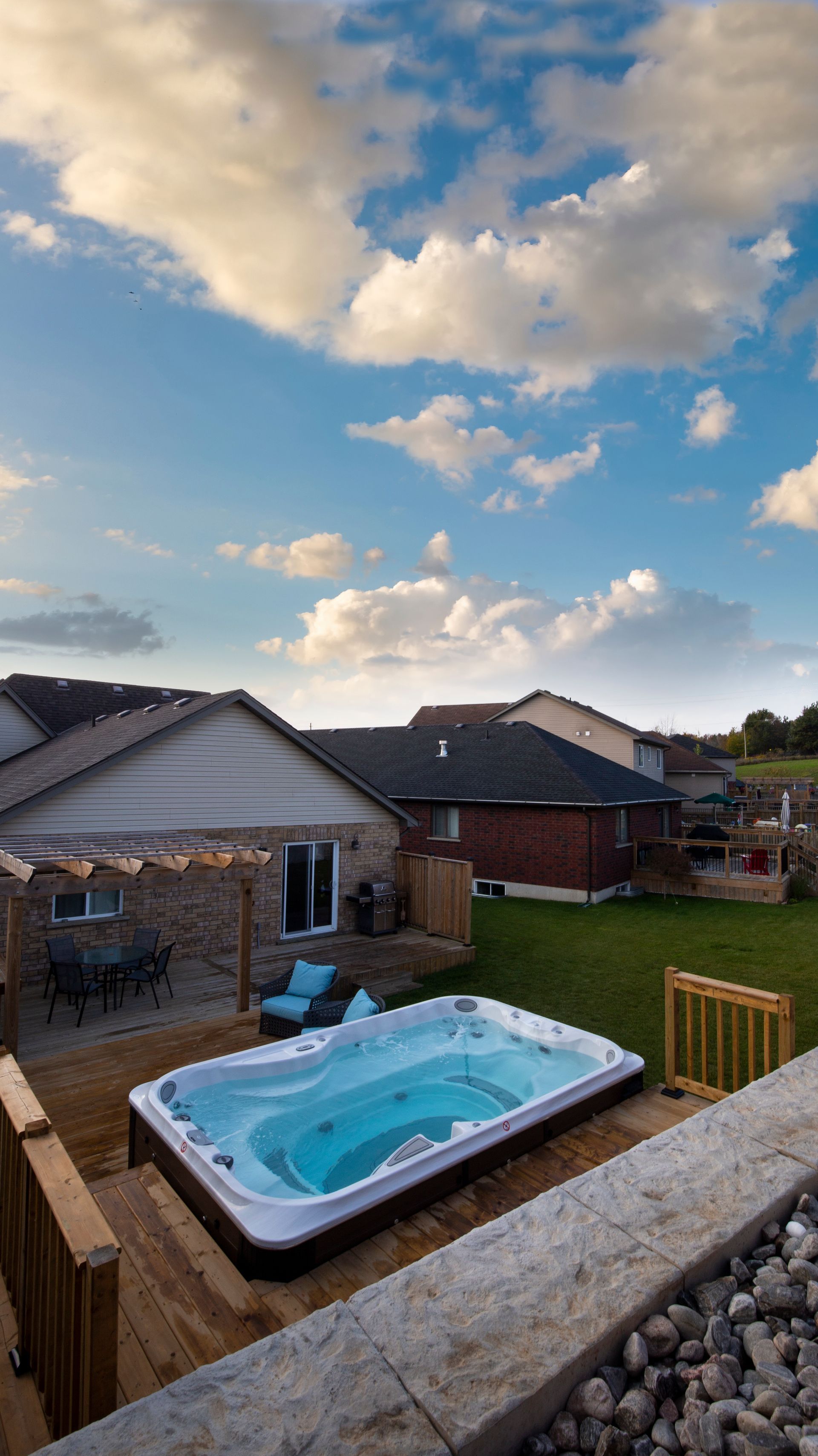 Jardin avec jacuzzi sur terrasse en bois, ciel bleu parsemé de nuages.