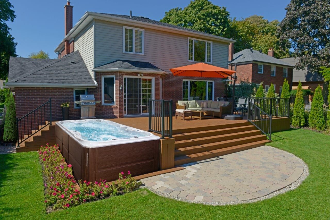 Jardin avec jacuzzi sur terrasse, mobilier de jardin et maison en briques, entouré de pelouse et d'arbres.