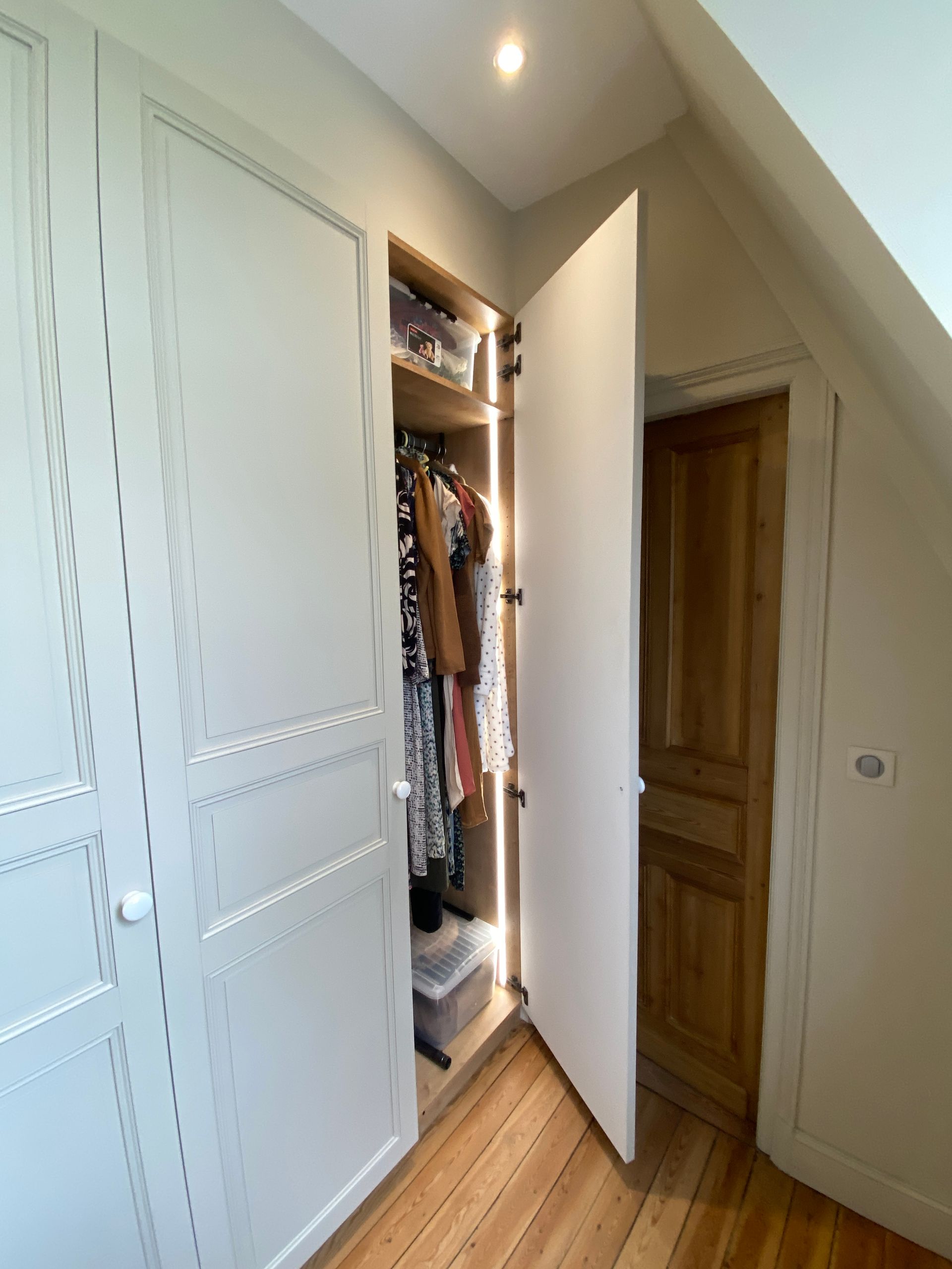 Placard intégré avec portes ouvertes donnant accès à des vêtements et à des espaces de rangement ; porte en bois à droite.