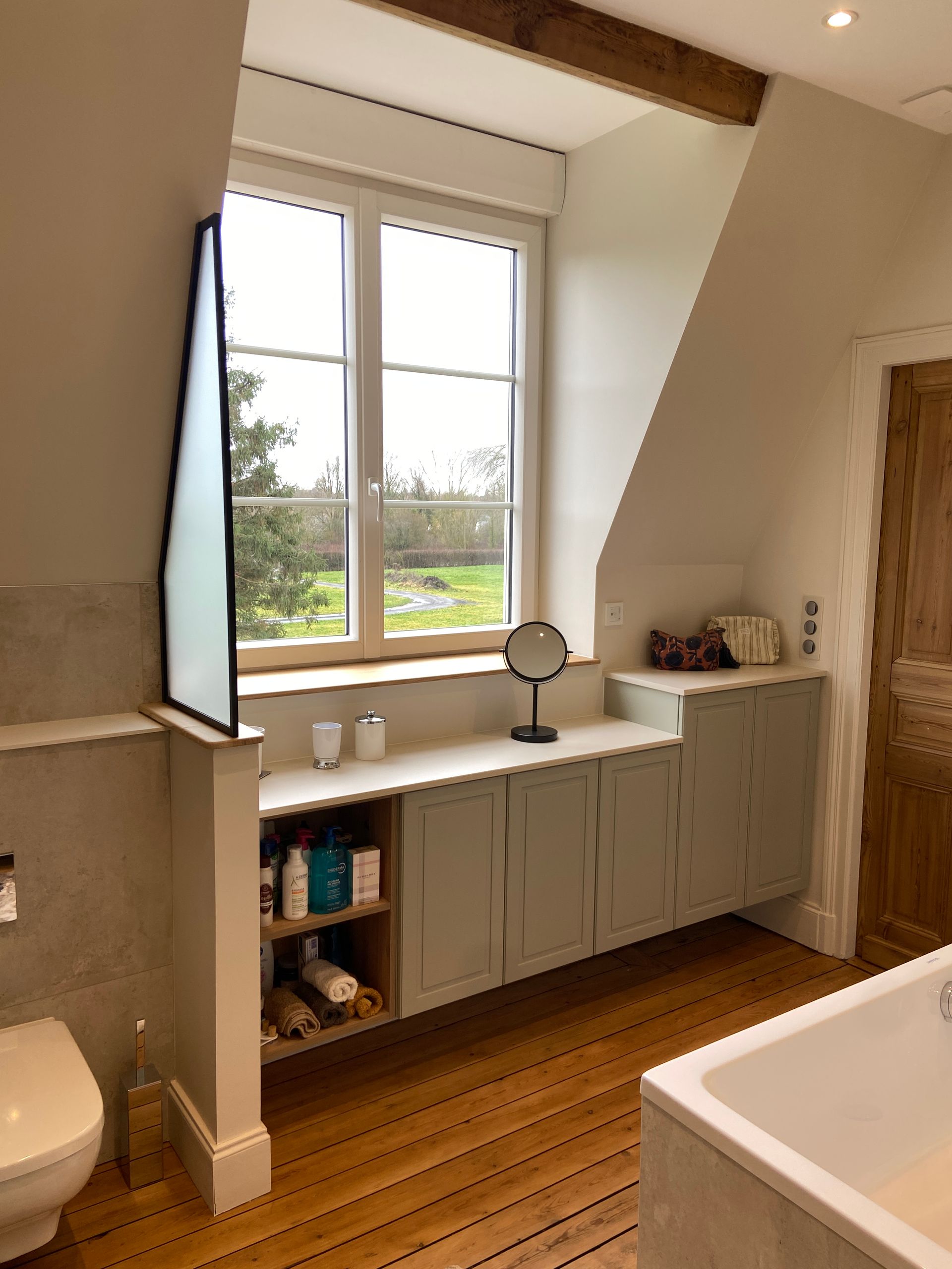 Salle de bains avec fenêtre, placards intégrés et parquet. Placards vert clair, murs blancs et vue sur un champ verdoyant.