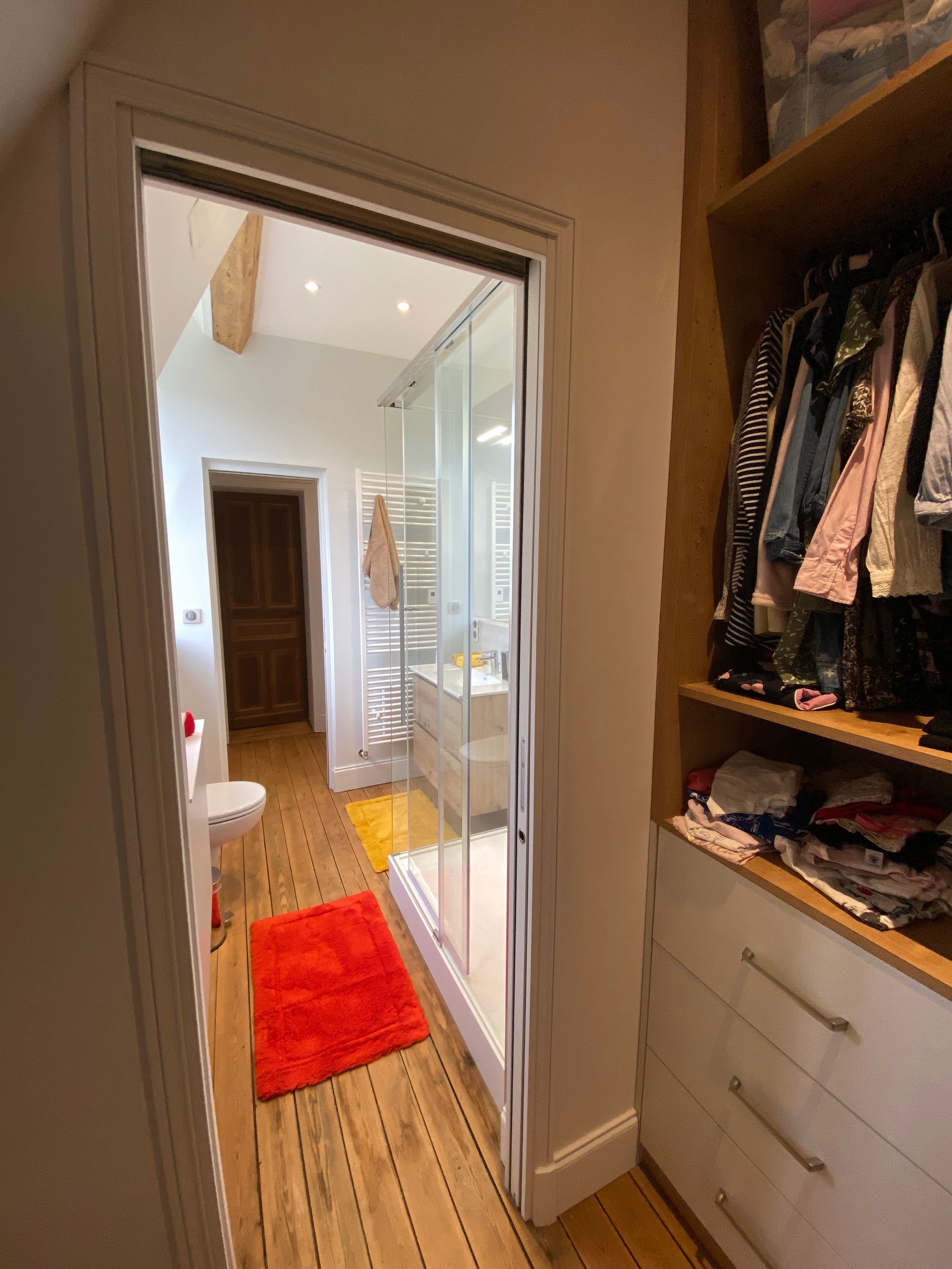 Dressing avec porte ouverte sur une petite salle de bains. Sol en bois, tapis orange, meubles blancs, vêtements et douche en verre.