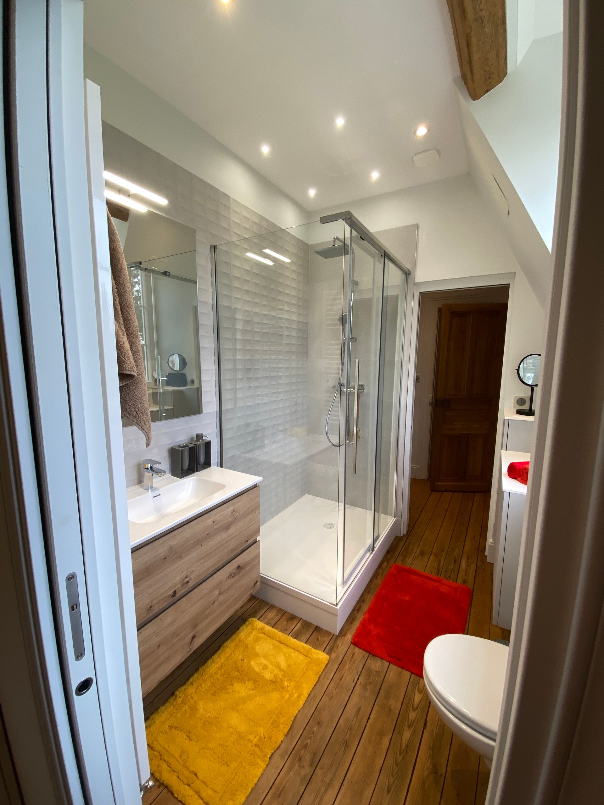 Salle de bains avec douche en verre, meuble-lavabo en bois, toilettes et deux tapis colorés sur parquet.