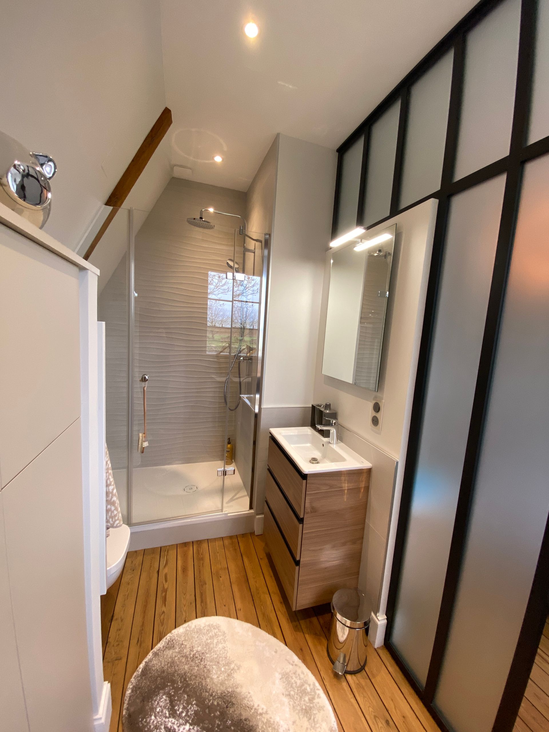Salle de bains avec parquet, douche en verre, meuble sous lavabo et porte en verre dépoli.