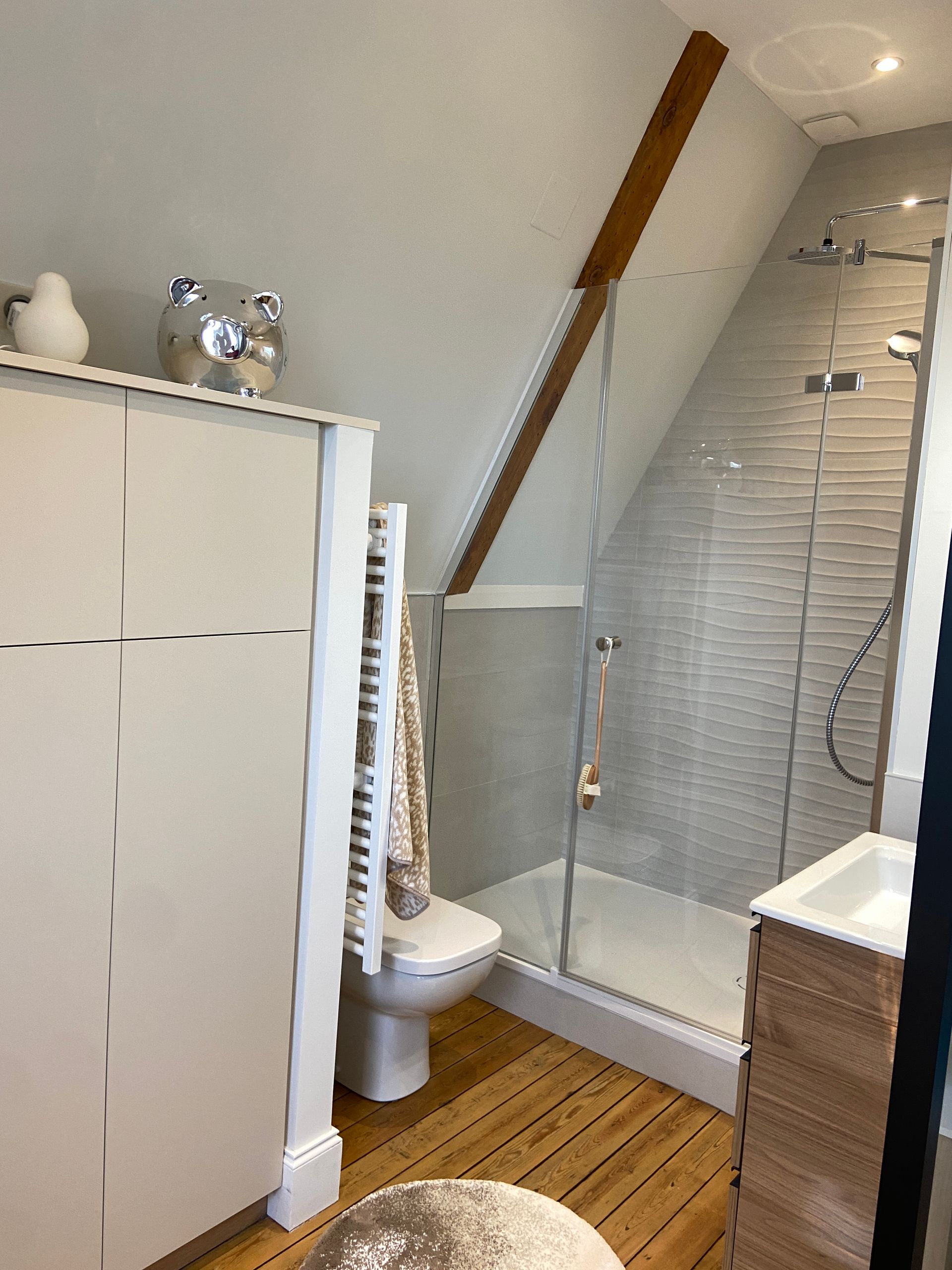 Salle de bains avec plafond mansardé, douche, toilettes, armoire, porte-serviettes et sol en bois.