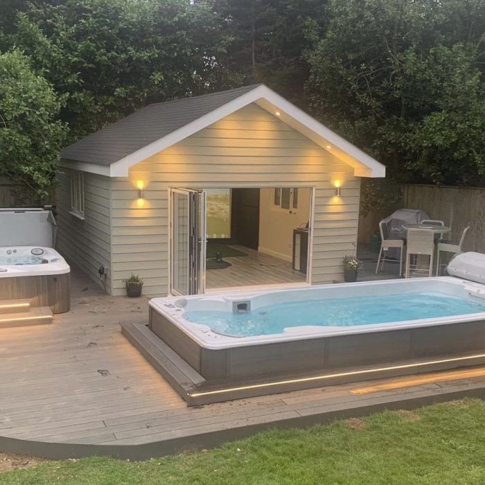 Un spa de nage, un jacuzzi et un petit bâtiment aux portes ouvertes, le tout entouré d'une terrasse en bois et d'aménagements paysagers.