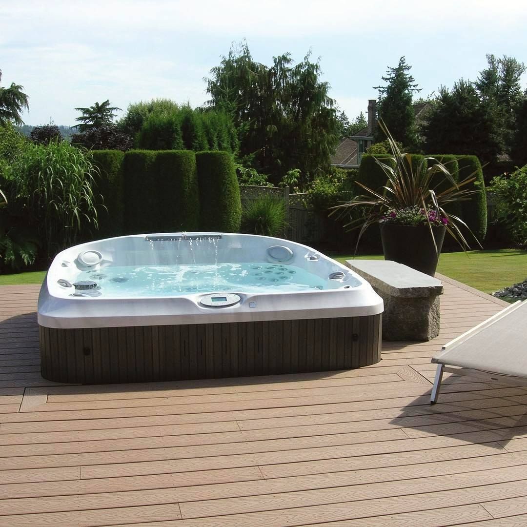 Un jacuzzi sur une terrasse en bois dans un jardin avec de la végétation luxuriante et un ciel d'un bleu limpide.