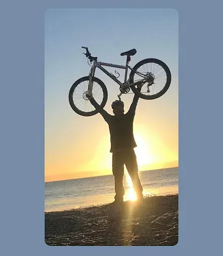Un hombre sostiene una bicicleta sobre su cabeza en la playa al atardecer.
