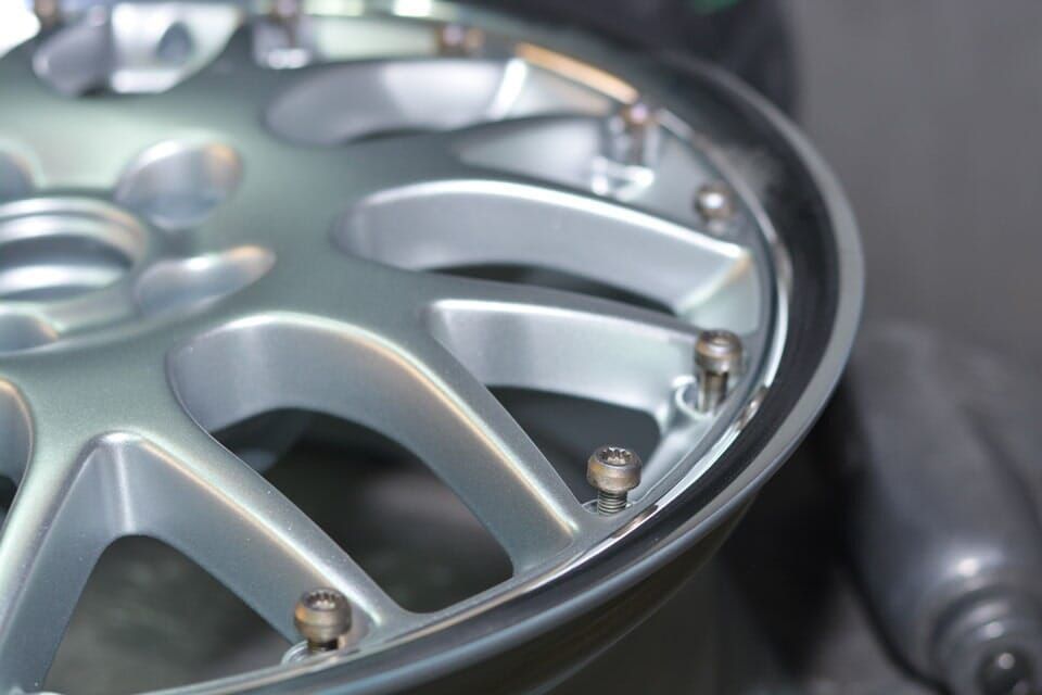 Roda de carro prateada com múltiplos raios, aro aparafusado, apoiada sobre uma superfície.