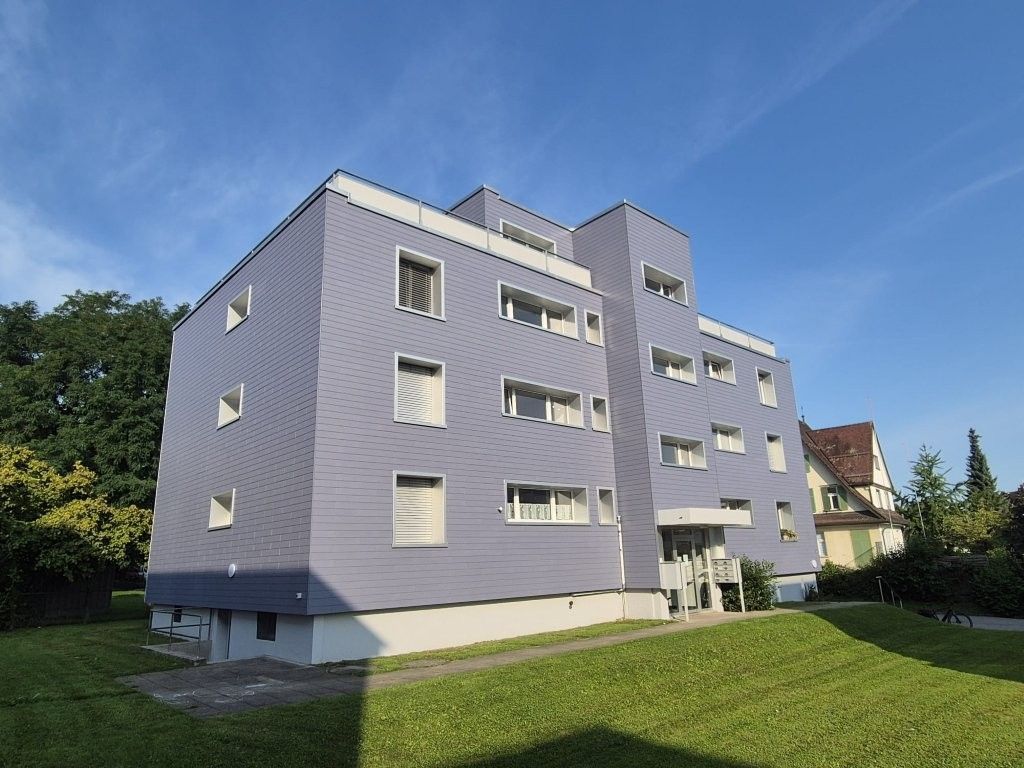 Modernes, grau-weißes Apartmentgebäude mit Balkonen, Bäumen und blauem Himmel.