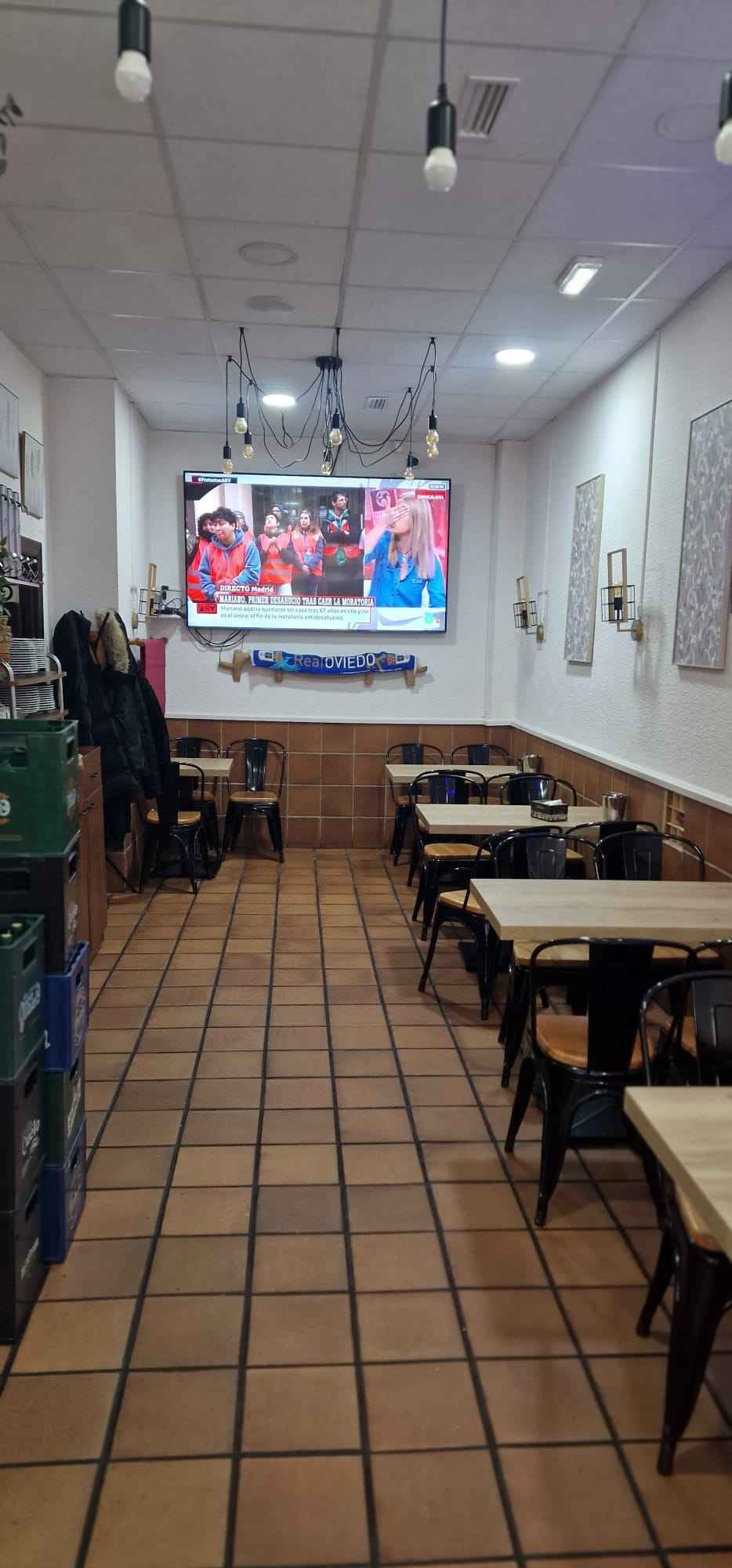 Interior de un restaurante con mesas y sillas, un televisor grande y luces colgantes.
