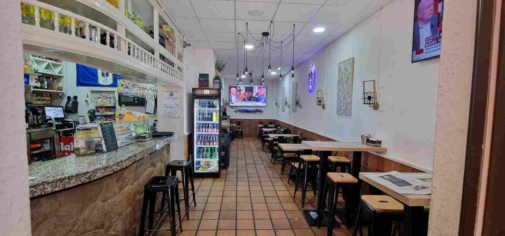Interior de un restaurante con barra, mesas y televisor. Paredes beige, suelo de baldosas y taburetes.