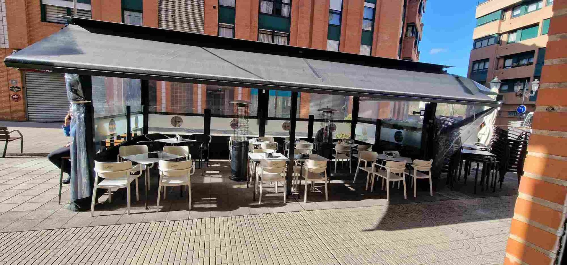 Restaurante al aire libre con mesas y sillas bajo un toldo. Edificios al fondo.