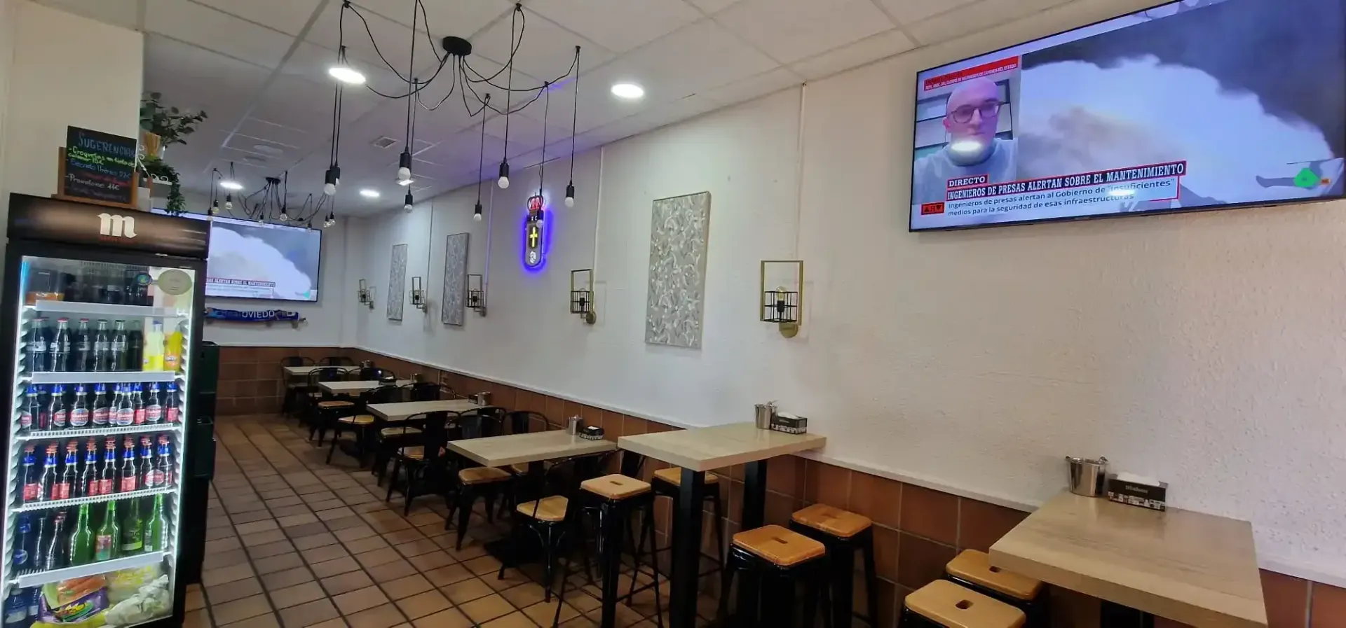 Interior de un restaurante con mesas, taburetes, televisores y un refrigerador lleno de bebidas.
