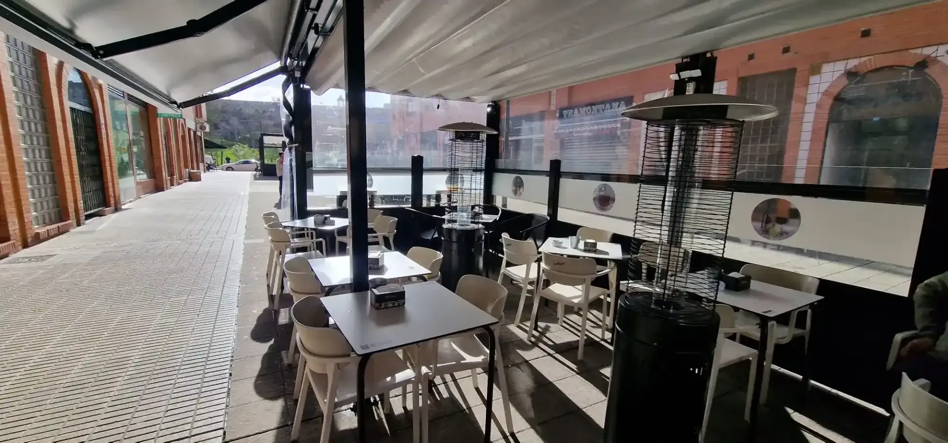 Una cafetería al aire libre con mesas, sillas y calefactores, cubierta por un toldo blanco. Un edificio de ladrillo al fondo.