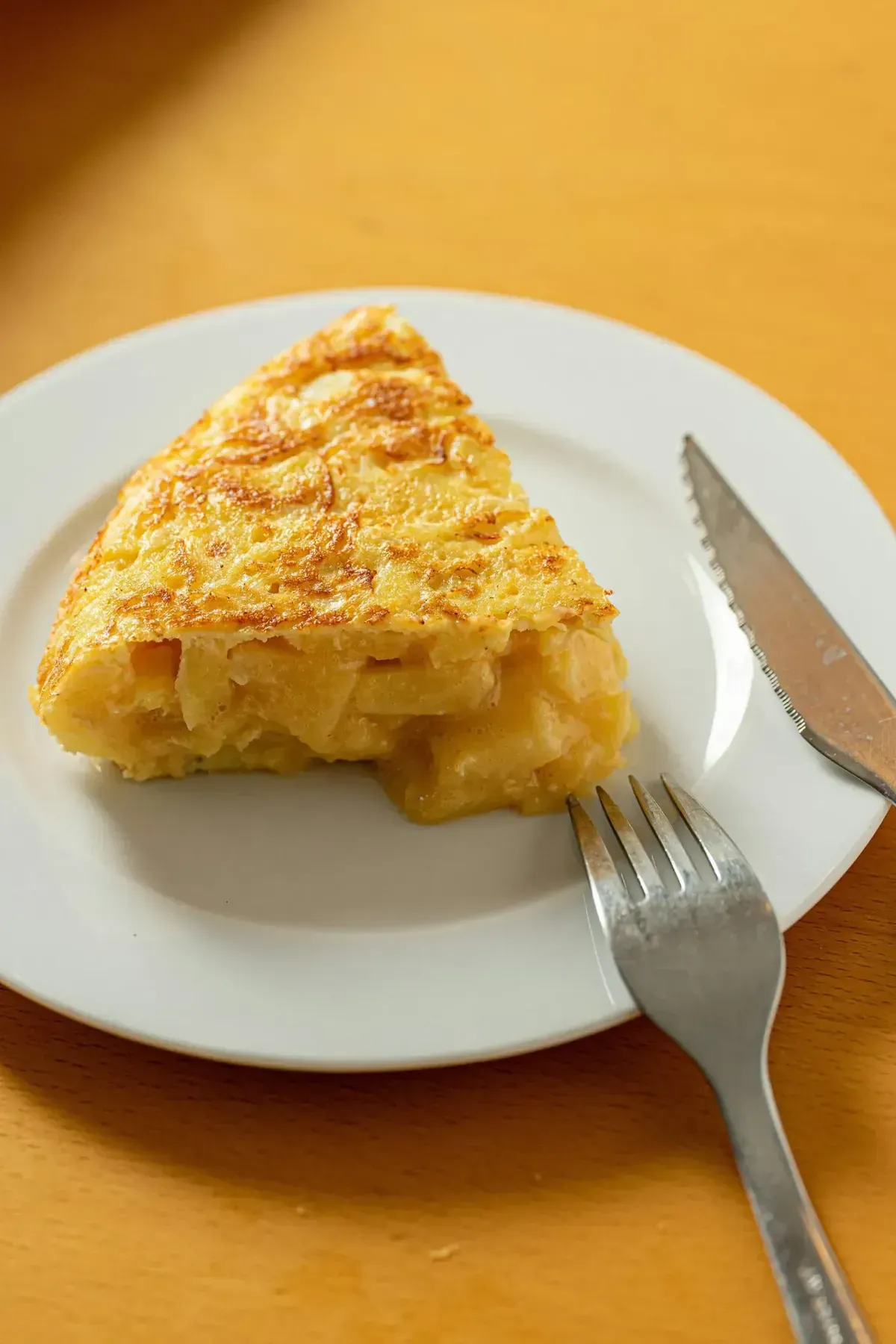 Rebanada de tortilla española en un plato blanco con un tenedor y un cuchillo sobre una superficie amarilla.