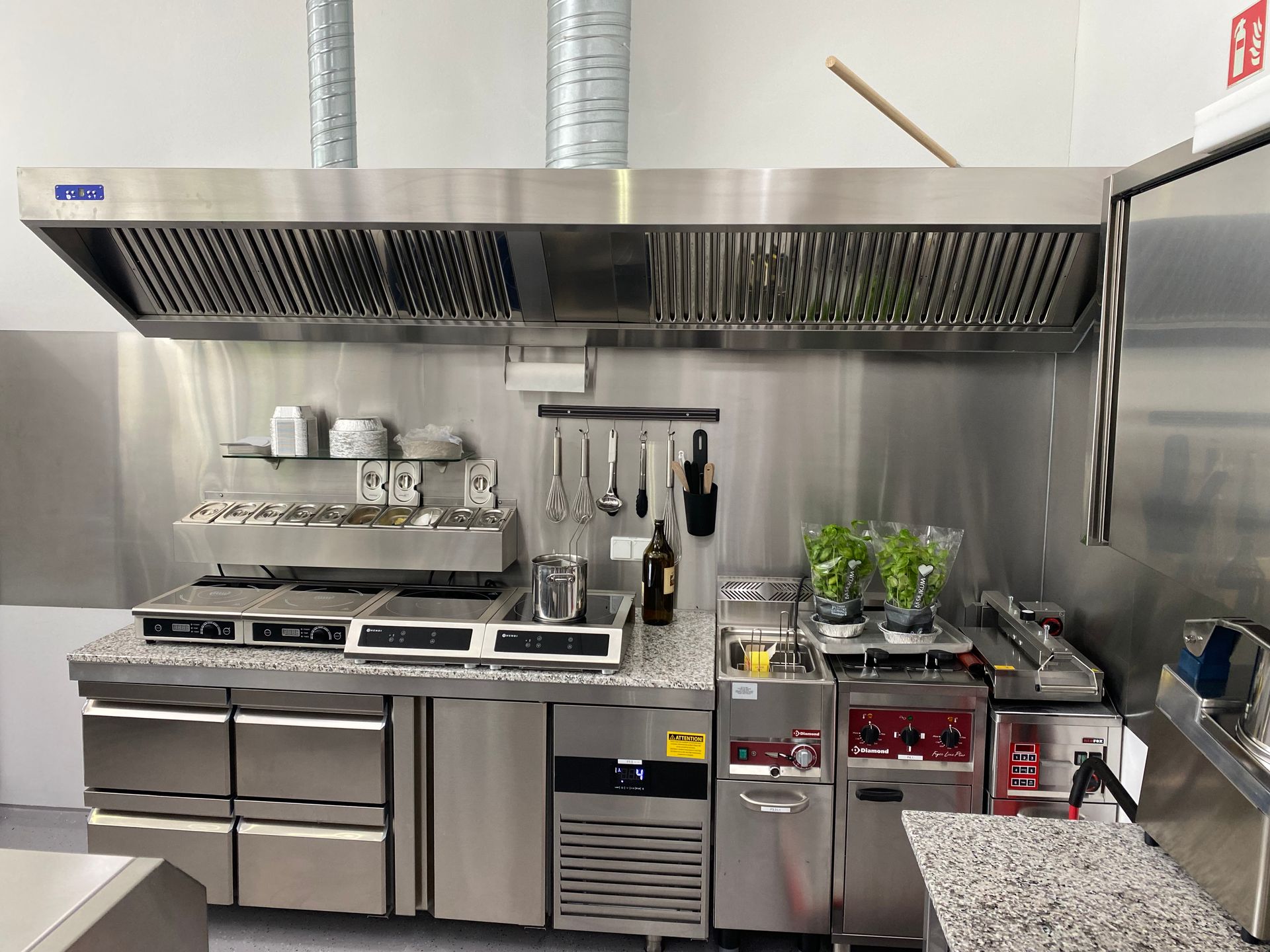 Professionelle Küchenvorbereitungsstation aus Edelstahl mit Arbeitsfläche, Spülbecken, Töpfen, Flaschen und Kräutern auf Regalen