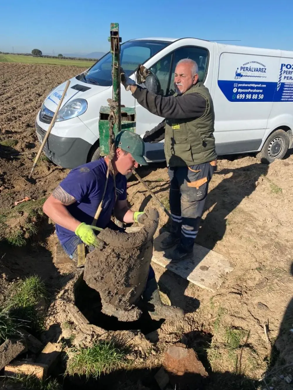 Dos personas trabajan con un objeto enterrado cerca de una camioneta en un campo. Una lo saca, otra maneja una herramienta.