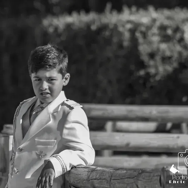 Un niño con traje blanco y corbata está apoyado contra una valla de madera.