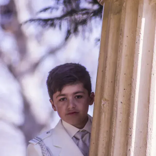 Un niño con traje y corbata está apoyado contra una columna.