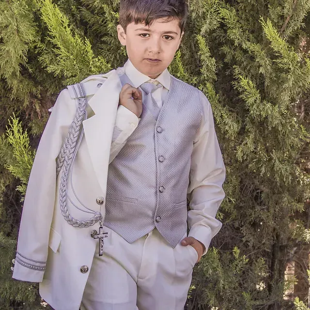 Un niño con traje y corbata está parado frente a unos árboles.