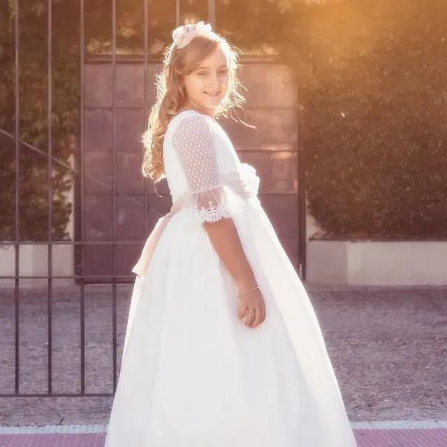 Una niña con un vestido blanco está parada frente a una valla.