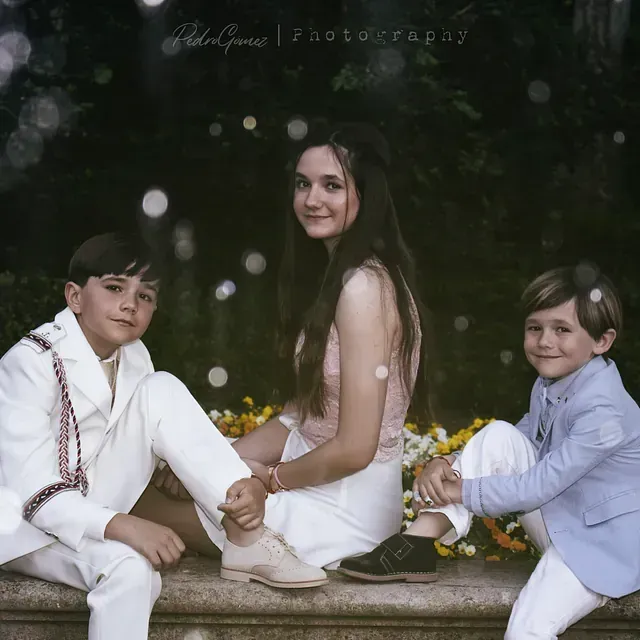 Una niña y dos niños están sentados en una repisa de piedra con flores al fondo.
